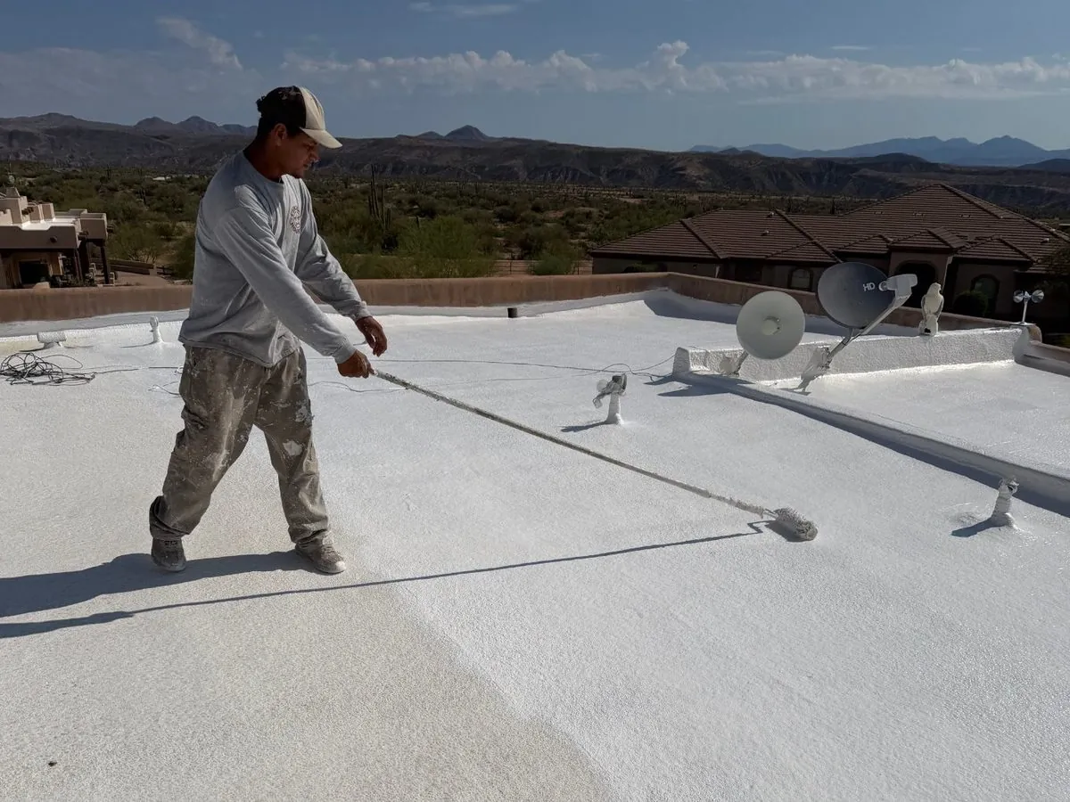 Crew member rolling protective coating across a finished white foam roof with the desert mountains behind
