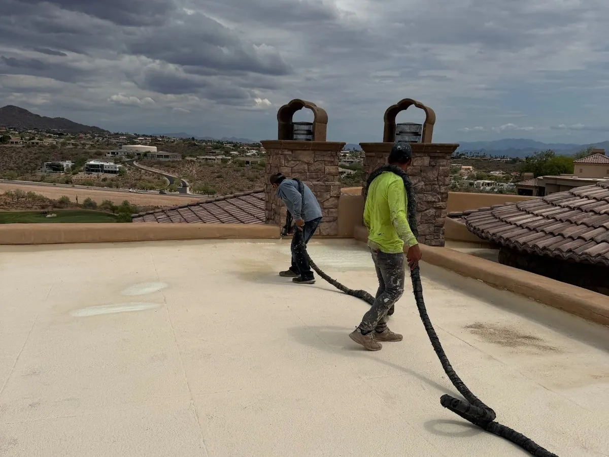 Two Arizona Tile Roofing crew members spraying foam on a flat roof as monsoon clouds build